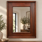 A rectangular wall mirror with a thick, dark brown wooden frame, styled on a wooden surface against a beige wall with decorative plants and a bowl.