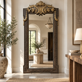 An ornate full-length floor mirror with a carved dark wood frame and gilded crest, standing in a bright, modern entryway.