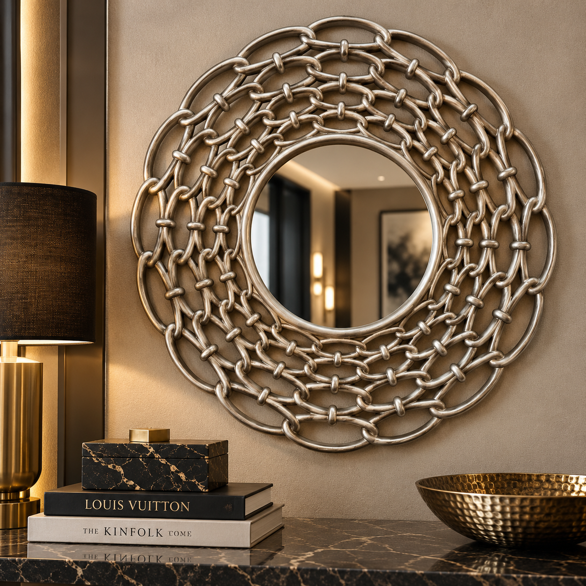 A large round decorative wall mirror with an intricate interlocking chain-link metal frame in a brushed silver finish, mounted on a beige wall above a marble console table with books and a lamp.