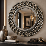 Round decorative wall mirror with an ornate chain-link style frame in distressed silver and dark gray, mounted on a beige wall above a console table with decor.