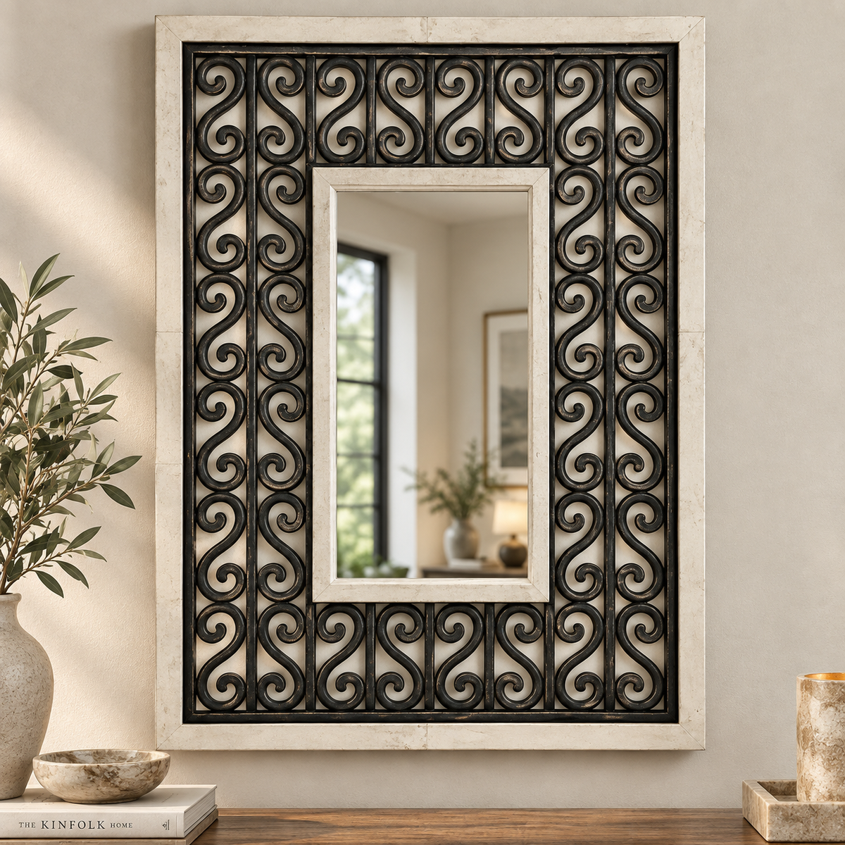 A rectangular decorative wall mirror with an ornate black scrollwork frame set within a light beige stone or plaster border, styled on a wooden surface against a beige wall with home decor props.