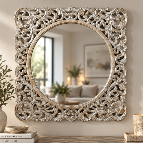 An ornate square wall mirror with a highly detailed, carved silver and gold baroque-style frame, shown hanging on a beige wall in a styled living room setting.