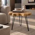 A live-edge wood accent table with black hairpin legs, styled in a modern living room next to a beige sofa with decorative books and a small ceramic vase on top.