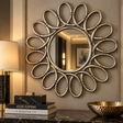 An ornate round wall mirror with a sculptural sunburst frame in a brushed silver finish, mounted on a beige wall above a dark marble console. A lit table lamp, stacked books including Louis Vuitton, and a hammered gold bowl are visible in the styled room setting.