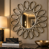An ornate round wall mirror with a sculptural sunburst frame in a brushed silver finish, mounted on a beige wall above a dark marble console. A lit table lamp, stacked books including Louis Vuitton, and a hammered gold bowl are visible in the styled room setting.