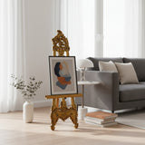 Luxury hand-carved wooden display easel featuring an ornate traditional design with intricate scrollwork and a rich antique gold finish, displayed with a framed abstract artwork. Ideal for showcasing artwork, mirrors, welcome signs, menus, or decorative displays in homes, galleries, boutique hotels, weddings, and event spaces.