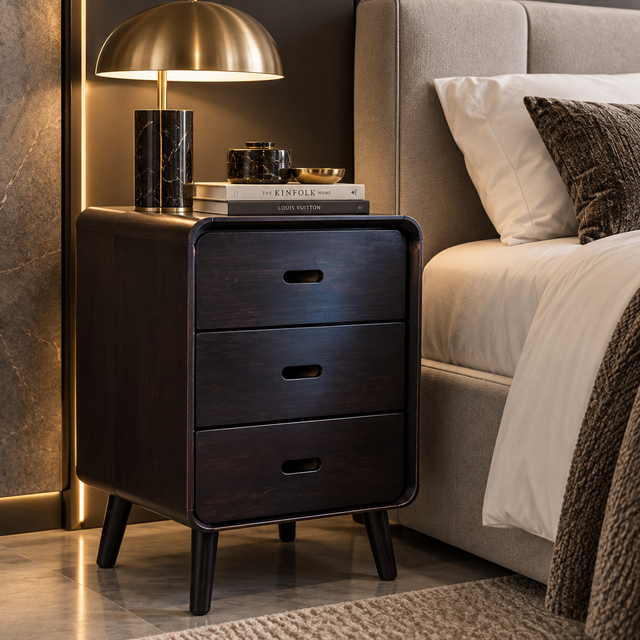 A dark wood three-drawer nightstand with tapered legs and recessed drawer pulls, styled beside a bed in a modern bedroom. A brass table lamp with a black marble base sits on top, along with stacked books and small decorative objects.