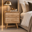 A mid-century modern wooden nightstand with two drawers and tapered legs, styled in a bedroom next to a bed with white linens and a brass table lamp on top.