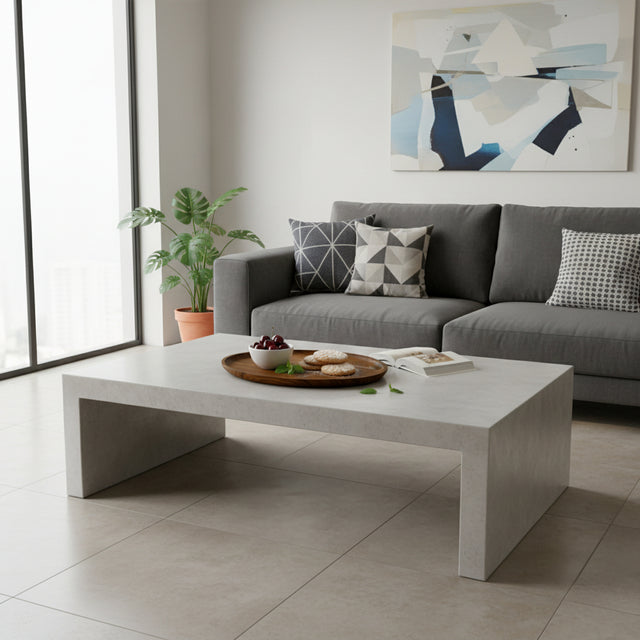 Modern minimalist rectangular coffee table with a light concrete-look finish and clean architectural lines, styled in a contemporary living room with a grey sofa and decorative tray.