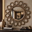 A large decorative wall mirror with an ornate sunburst frame made of interlocking circular wood elements, mounted on a beige wall above a dark marble console. The reflection shows a dimly lit interior, and the surrounding decor includes a metallic vase, a bowl, books, and sculptural objects.