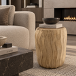 A natural wood barrel-shaped side table with wavy grain patterns, styled in a modern living room next to a beige textured sofa and a fireplace.