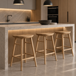 Three backless natural wood bar stools with curved saddle seats, splayed legs, and wooden footrests beside a kitchen island.