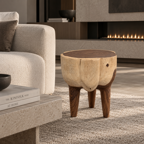 A rustic wooden stool with a natural log top and three sturdy legs, styled in a modern living room next to a beige armchair and in front of a fireplace.