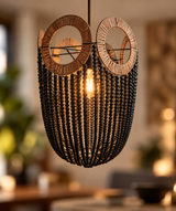 A bohemian-style pendant light fixture featuring a black beaded basket body, two circular woven wood sunburst rings, and a glowing warm bulb in the center, shown hanging in a softly blurred indoor setting.