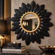 A decorative wall mirror with a sunburst leaf design in black and a gold-toned circular frame, mounted on a beige wall above a console table with books and a bowl.