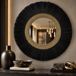 A large round wall mirror with a black fringed outer border and a woven beige inner ring, mounted on a gray wall above a console table with decorative vases, a bowl, and sculptural objects.