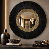 A large round wall mirror with a black fringed outer border and a woven beige inner ring, mounted on a gray wall above a console table with decorative vases, a bowl, and sculptural objects.