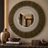 A large round decorative wall mirror with an ornate white geometric pattern border and a dark olive green fringed sunburst edge, mounted on a beige wall above a marble console with a gold vase and books.