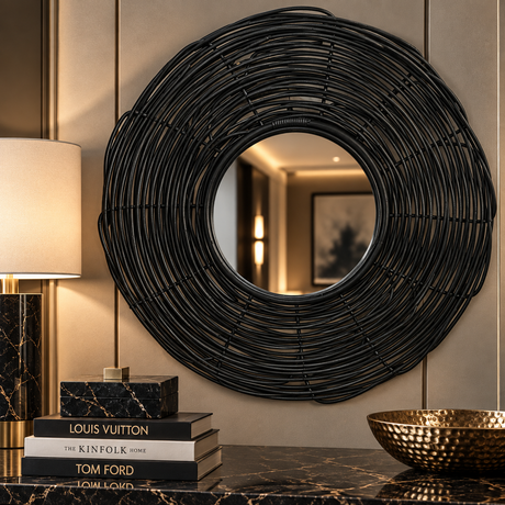 A large circular black woven wall mirror with a thick coiled frame, mounted on a beige paneled wall above a dark marble console table with books and a gold bowl.