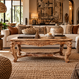 An ornate carved wooden coffee table in a rustic, bohemian-style living room. The table is styled with books, a ceramic vase, and decorative bowls on top, set on a large woven jute rug in front of a cream-colored sofa with patterned throw pillows.
