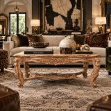 An ornate carved wooden coffee table in a styled living room setting, featuring a beige sofa, leopard print and patterned throw pillows, a large framed map artwork on the wall, and decorative objects on the tabletop.