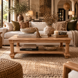 A rustic wooden coffee table with a double shelf sits in a beautifully styled living room. The table is styled with books, a candle, and decorative ceramic vases containing dried botanicals.