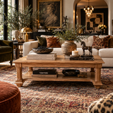 A styled rustic wooden coffee table in a richly decorated living room, featuring books, a bowl of blueberries, a ceramic vase with olive branches, a small horse figurine, and a candle on its shelves.