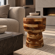 A rustic wooden side table with a chunky, stacked natural wood grain finish stands in a modern living room next to a beige sofa and a dark marble coffee table.