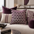 A styled living room scene showing a beige textured sofa with multiple decorative throw pillows. The central pillow features a dark purple polka dot pattern on a cream background, surrounded by other purple and beige patterned pillows.