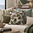 A styled living room scene with multiple decorative throw pillows on a light upholstered sofa. The central pillow features a cream background with a botanical print in olive green, brown, and muted blue, surrounded by coordinating pillows in striped, olive checkered, solid sage, and textured beige.