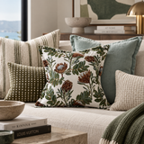 A styled living room scene with multiple decorative throw pillows on a light upholstered sofa. The central pillow features a cream background with a botanical print in olive green, brown, and muted blue, surrounded by coordinating pillows in striped, olive checkered, solid sage, and textured beige.