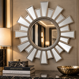 A round sunburst wall mirror with a silver-toned beaded frame and radiating rectangular mirror segments, mounted on a beige wall above a dark marble surface with decorative books, a lamp, and a gold bowl.