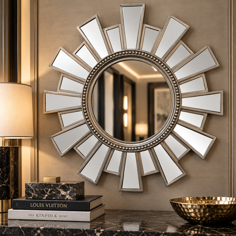 A round sunburst wall mirror with a silver-toned beaded frame and radiating rectangular mirror segments, mounted on a beige wall above a dark marble surface with decorative books, a lamp, and a gold bowl.