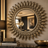 An ornate round wall mirror with a sunburst leaf design frame in a distressed gold finish, mounted on a beige wall above a marble console table with decorative books and a lamp.