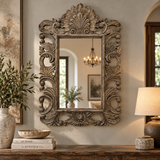 An ornate, vintage-style rectangular wall mirror with a heavily carved distressed wood frame, mounted on a beige wall in a styled home interior above a wooden console table.