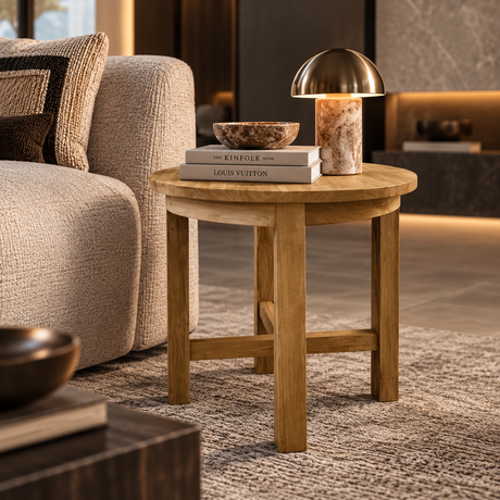 A round wooden end table in a warm beige tone, styled in a modern living room next to a textured sofa. On top of the table are two books, a small bowl, and a table lamp with a metallic dome shade and a marble base.