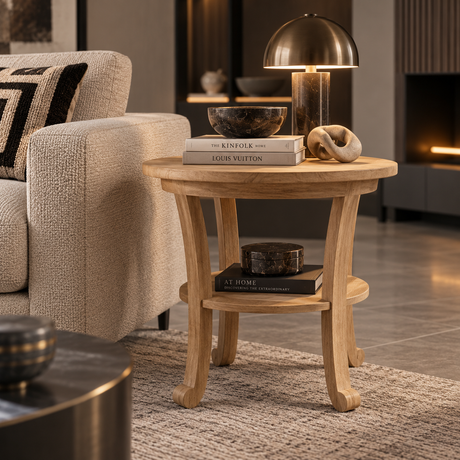 A round light oak end table with a lower shelf, styled in a modern living room setting with a brass dome lamp, books, and decorative bowls.