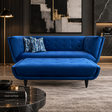 A modern blue velvet chaise lounge with a matching ottoman in a styled living room setting. The lounge features tufted detailing on the backrest and seat, tapered black legs, and is styled with two blue patterned throw pillows.