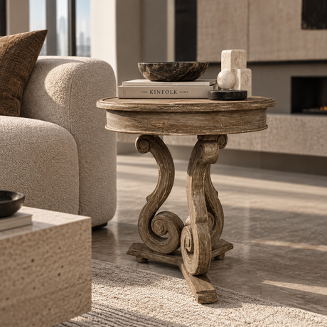 A round rustic wooden side table with ornate carved legs, styled in a modern living room next to a beige armchair.