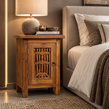 A rustic wooden nightstand with carved details and a single cabinet door, styled next to a bed in a warm, modern bedroom setting.