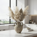 Rustic ceramic amphora vase with a textured stone finish and twin loop handles, styled with pampas grass, decorative branches, and natural botanicals on a modern coffee table, adding timeless organic elegance to contemporary interiors.