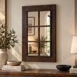 A dark brown wooden wall mirror with a six-pane grid design, styled on a wooden console table against a beige wall with decorative books, a plant, and a lamp.