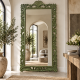 An ornate full-length floor mirror with a carved green frame stands in a bright, modern room. The mirror reflects an arched doorway and a living room interior, while the surrounding environment features a wooden console table, a vase of white flowers, and a large potted olive branch.
