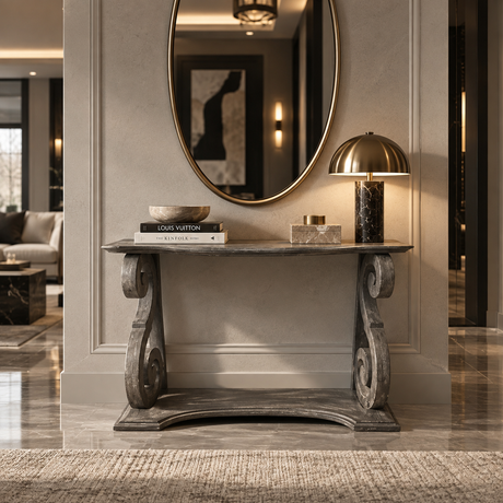 A dark gray carved console table in a luxury entryway, styled with books, a marble bowl, a small box, and a gold dome lamp. An oval gold-framed mirror hangs on the wall behind it.
