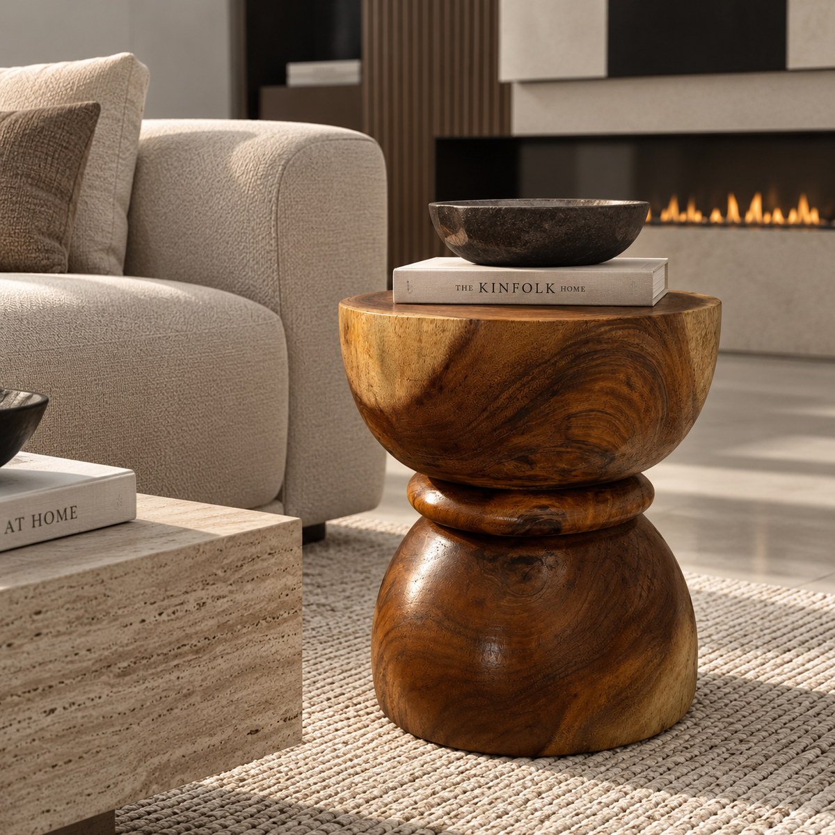 A round wooden side table with a carved pedestal base and a wide top, styled in a modern living room next to a beige sofa and in front of a fireplace.
