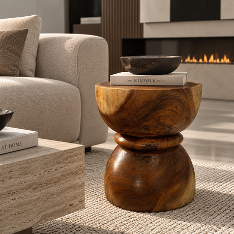 A round wooden side table with a carved pedestal base and a wide top, styled in a modern living room next to a beige sofa and in front of a fireplace.