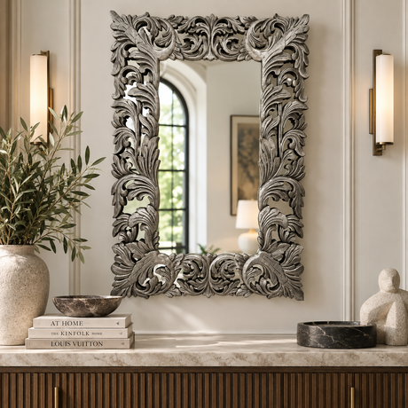 An ornate rectangular wall mirror with a highly detailed, carved silver-gray floral and scrollwork frame, displayed in a styled interior setting above a console table.