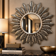 A round decorative wall mirror with a sunburst leaf design in a distressed silver and gold finish, mounted on a beige wall above a dark marble console table with books and a gold bowl.