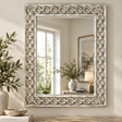 A large rectangular wall mirror with an ornate distressed silver frame featuring interlocking circular motifs, styled on a wooden table next to a vase with greenery.