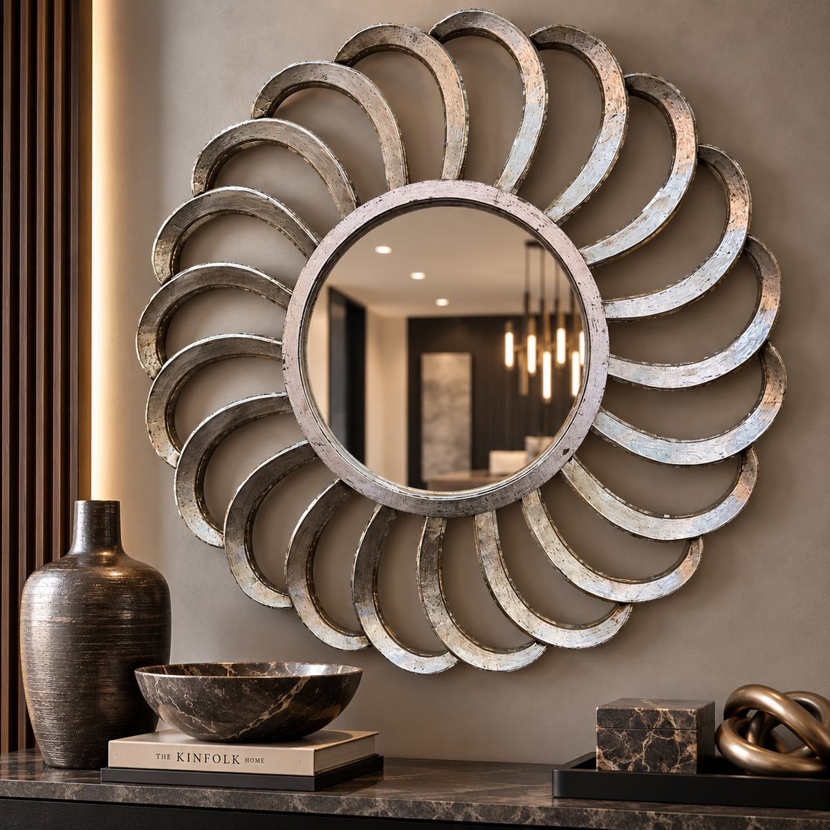 A large decorative wall mirror with a sunburst frame made of overlapping metallic rings, mounted on a taupe wall above a console table with vases and decor.
